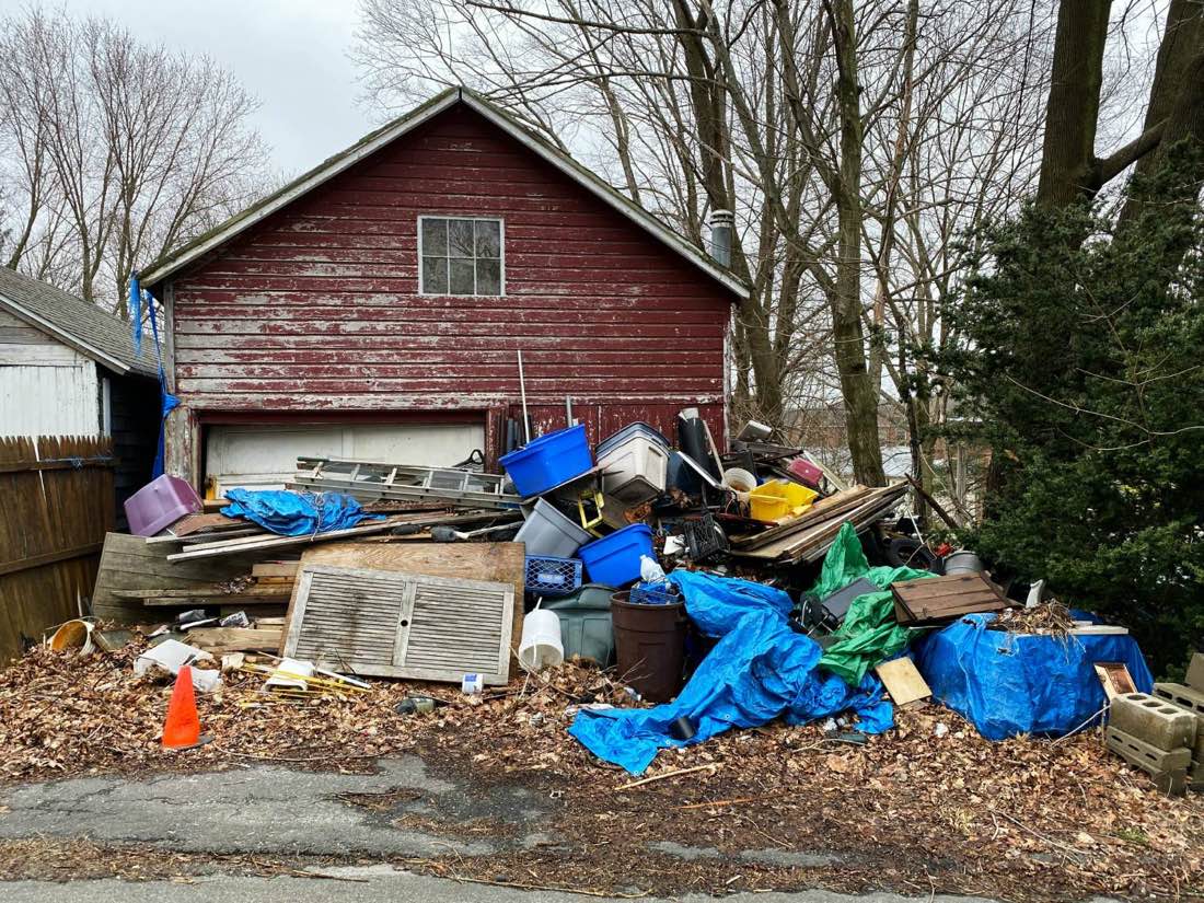 Large pile of dumped debris, lumber, bins, and tarps in front of an old garage, waiting to be hauled off